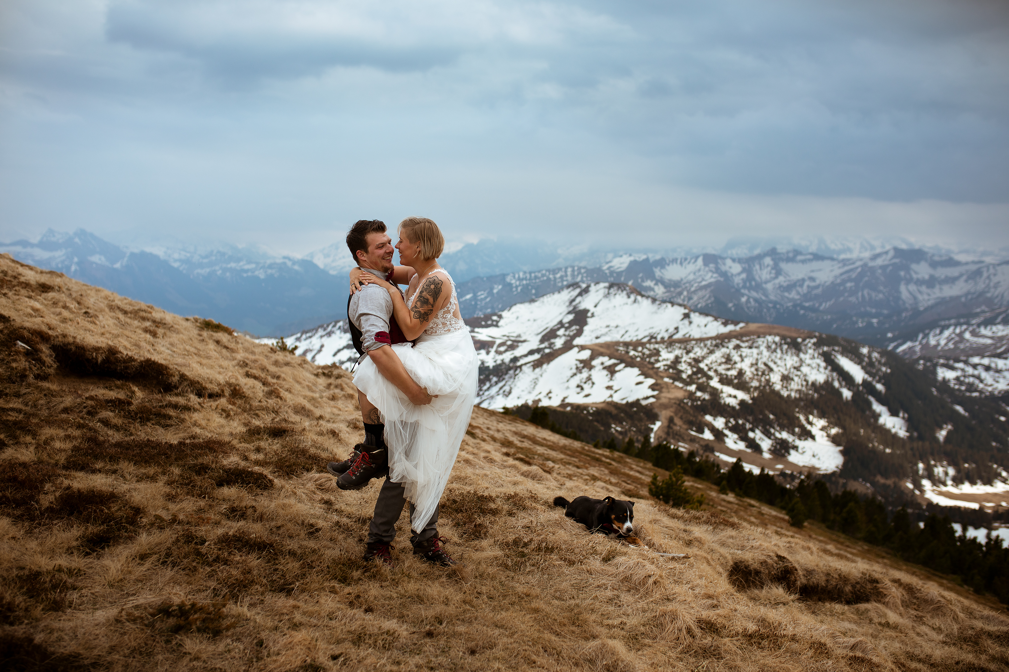 Das Brautpaar hat sich für eine Hochzeit zu Zweit in den Schweizer Bergen entschieden. Braut und Bräutigam tragen Hochzeitskleid, Anzug und Wanderschuhe. Die Trauung findet auf dem Rickhubel beim Glaubenbergpass bei Sarnen statt. Der Hund der Beiden - ein Appenzellermix ist bei der Hochzeit dabei, und kaut während dem Brautpaarshooting gemütlich auf einem Stock.